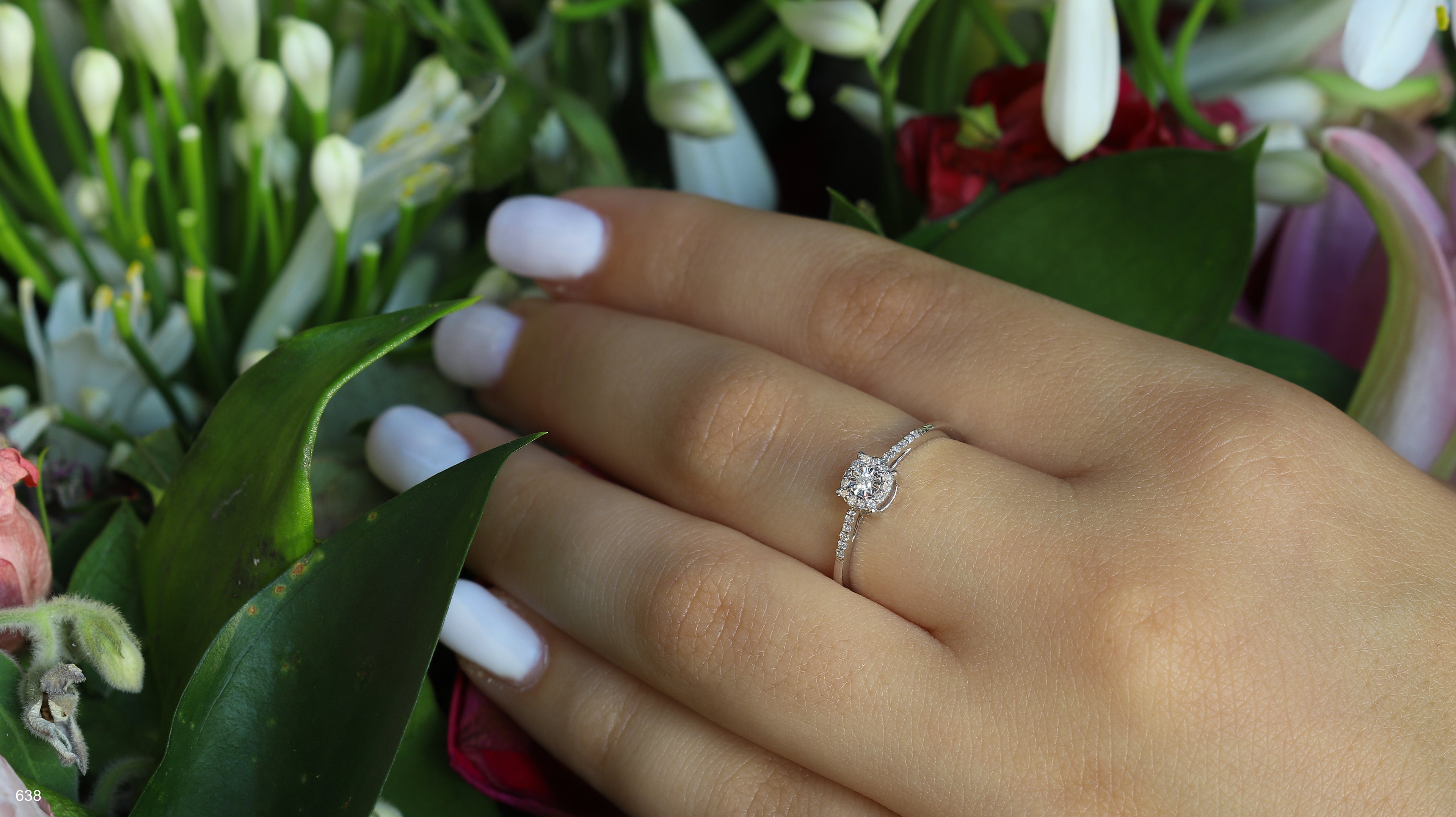 יד עם ציפורניים לבנות מטופחות טריות לובשת טבעת עדינה, מוקפת בפרחים צבעוניים.